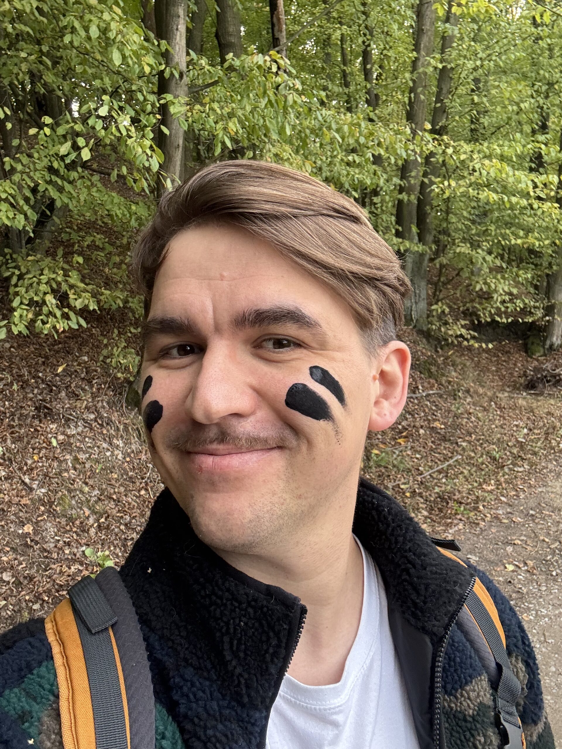 A smiling person standing in a forest with black face paint markings on their cheeks, wearing a backpack and outdoor clothing.