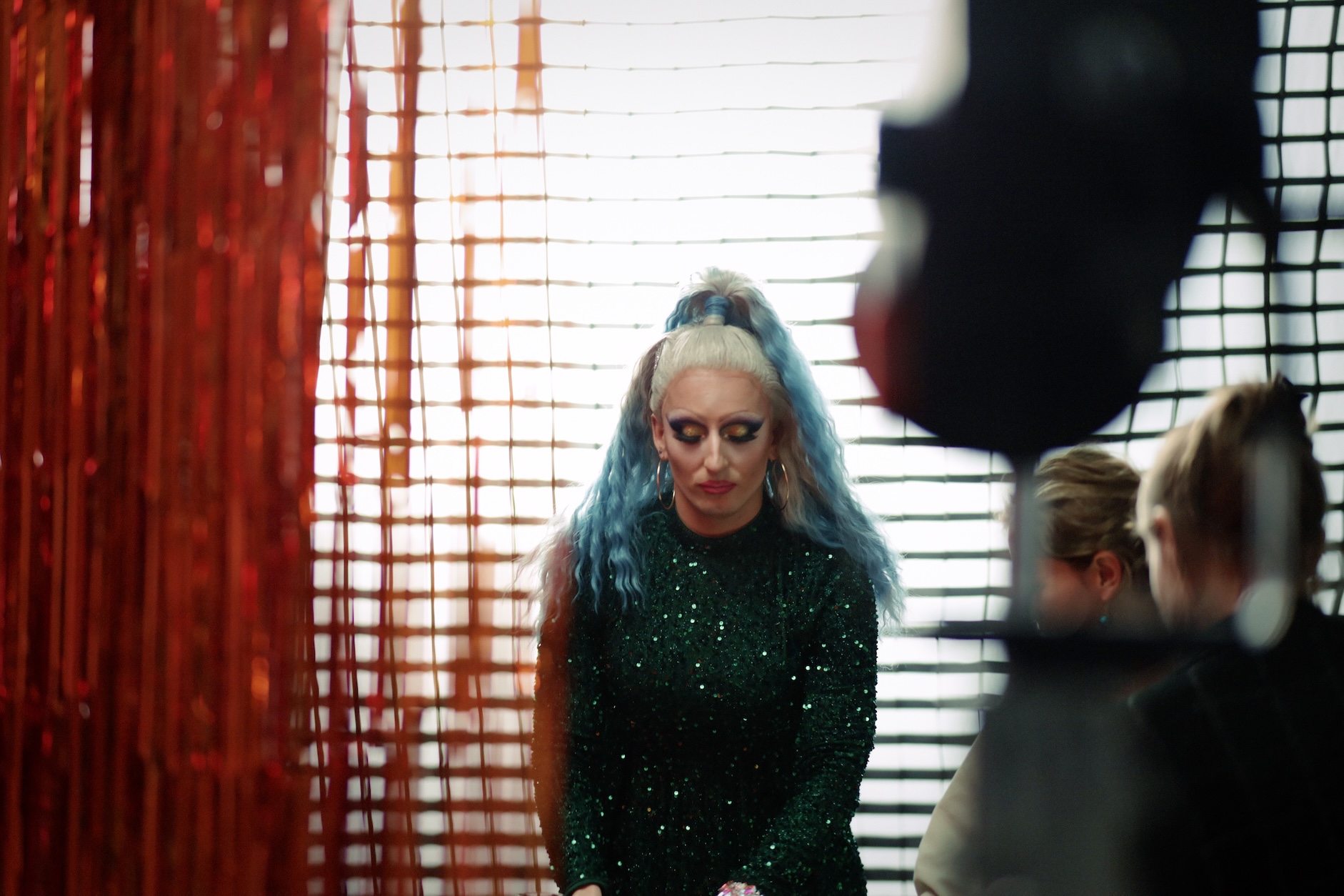 Drag performer w błyszczącym zielonym stroju i turkusowych włosach stoi na planie zdjęciowym z czerwoną wstążkową dekoracją w tle