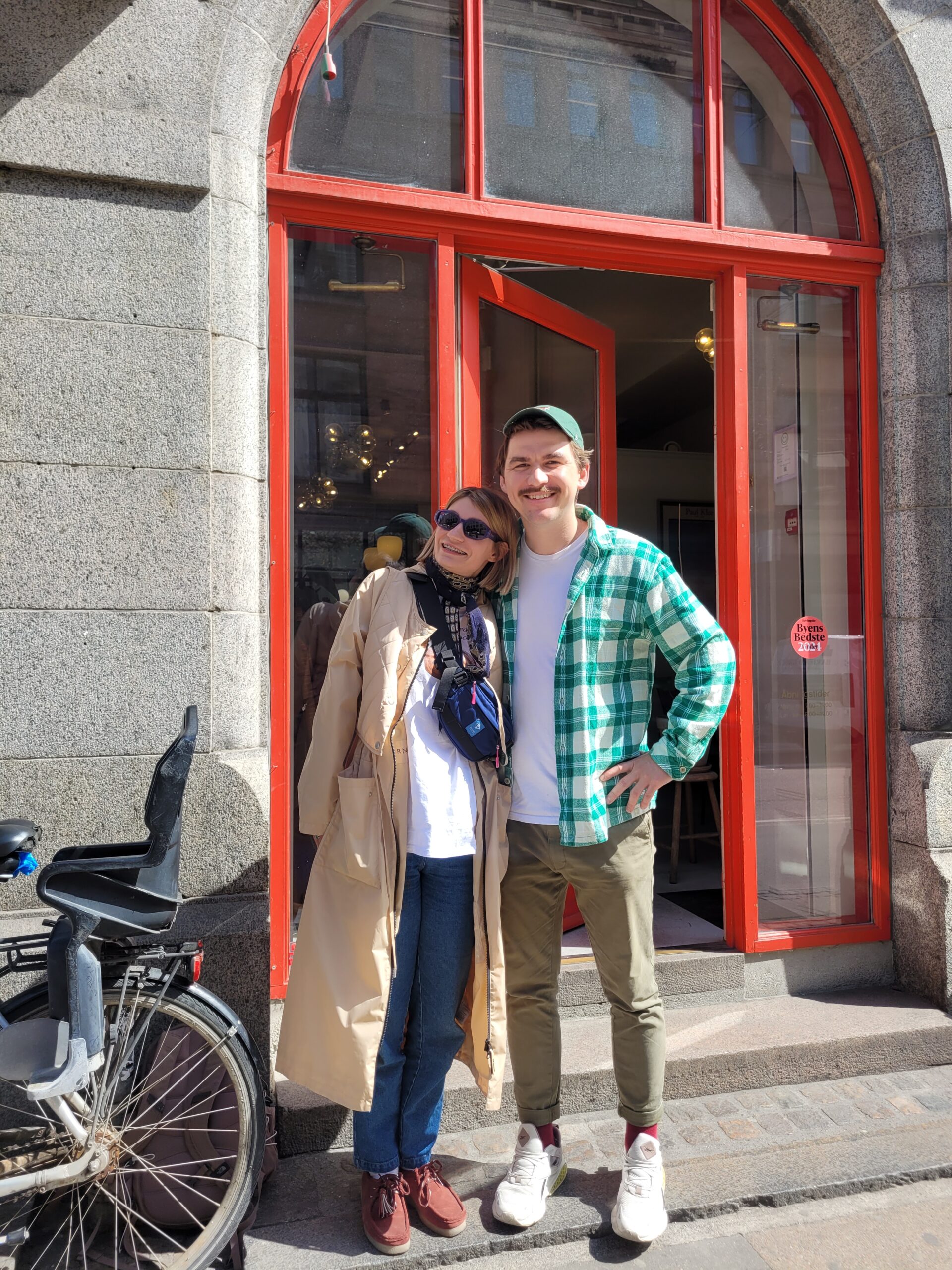 Two people standing close together and smiling in front of a building with a large red-framed doorway; one wearing a long beige coat and sunglasses, the other in a green checkered shirt and cap, with a bicycle visible on the left side.