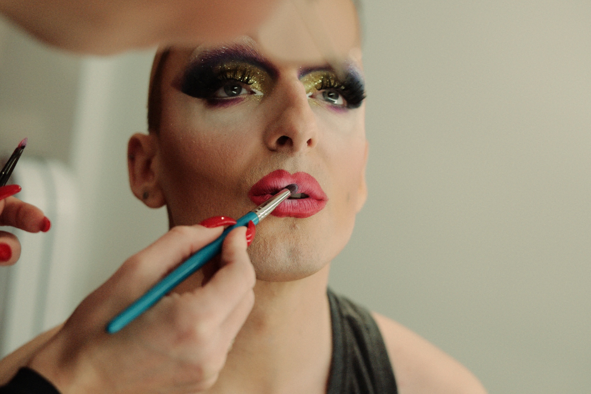 Zbliżenie na aplikowanie makijażu drag - pędzelek nakłada czerwoną szminkę na usta, w tle widoczny dramatyczny makijaż oczu ze złotymi akcentami