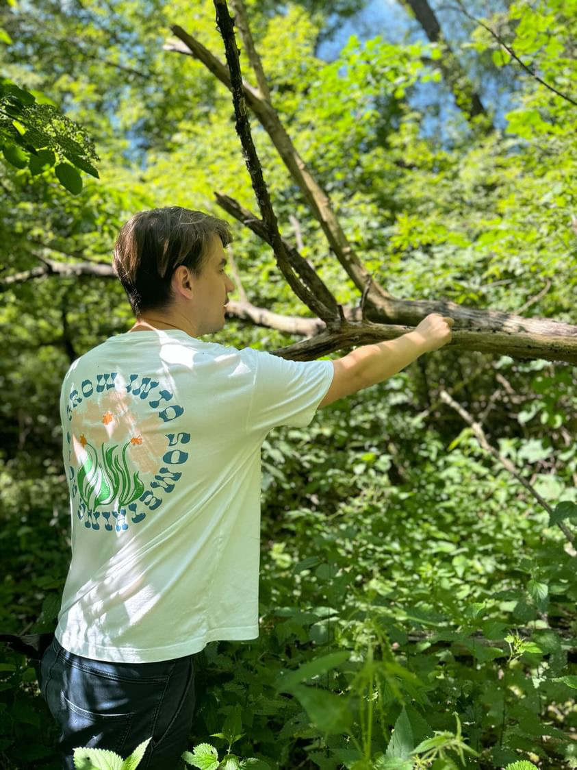 A person standing in dense forest greenery, holding a large branch and looking into the woods, wearing a white T-shirt with a graphic print on the back.